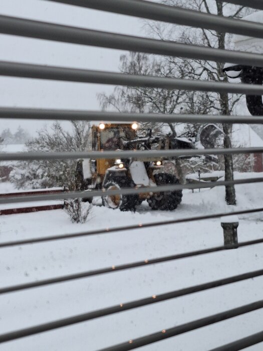 En snöplogmaskin plogar en snötäckt uppfart, sedd genom persienner. Träden i bakgrunden är snötäckta.