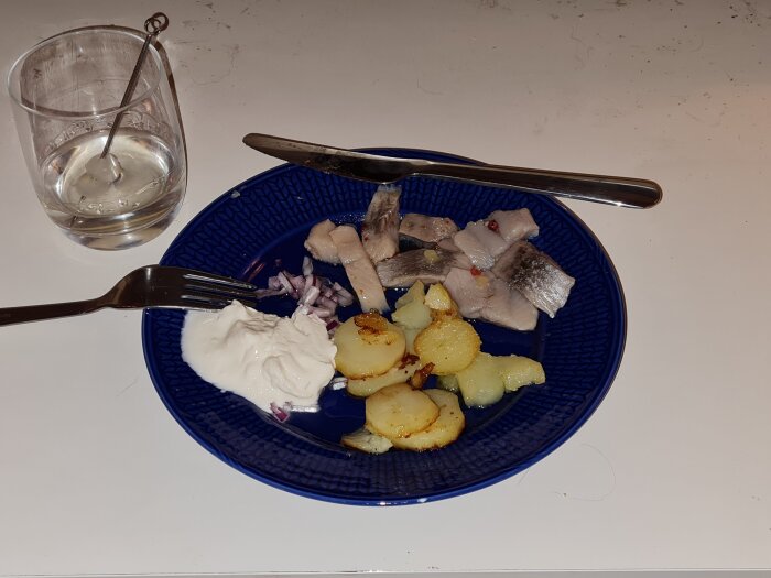 Blå tallrik med sillbitar, stekt potatis och gräddfil. Bredvid tallriken finns ett glas med en cocktailpinne.