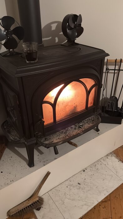 Vedeldad kamin med smutsigt glas och sotig yta, omgiven av askrester och borste.