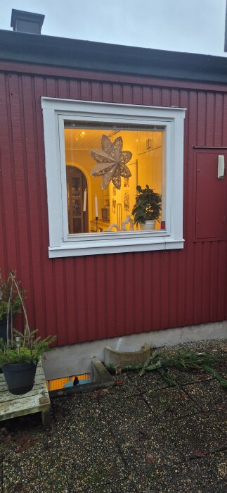 Rött hus med vitt fönster, genom vilket en stjärnformad dekoration och en krukväxt syns inomhus.