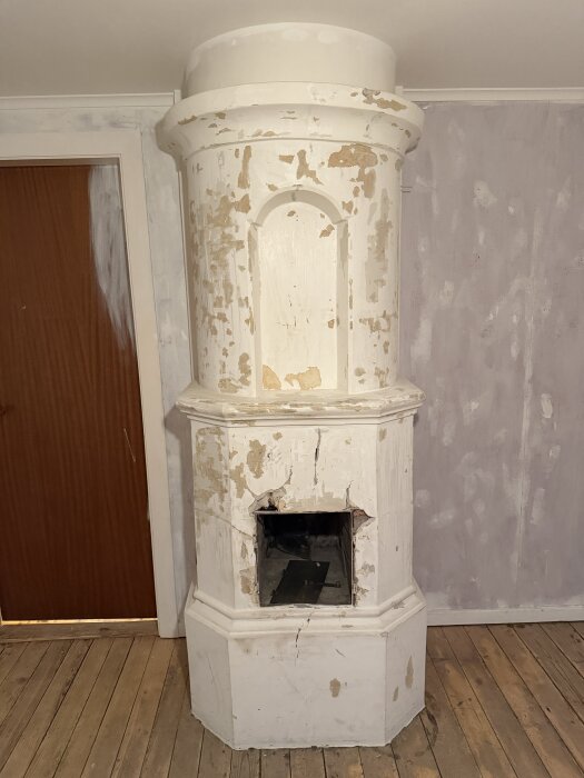Rörspis i behov av renovering, med avskavd färg och sprickor, visar exponerade ytor och öppning mot äldre kakelugn i ett hus.