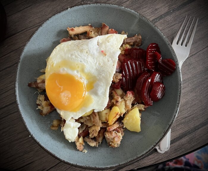 Pyttipanna med stekt ägg och inlagda rödbetor på en tallrik, serverad på ett bord.