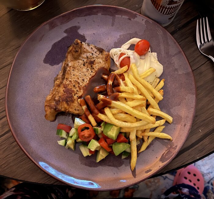 Fläskkotlett med pommes frites, avokado- och tomatsallad och en klick sås på tallrik.