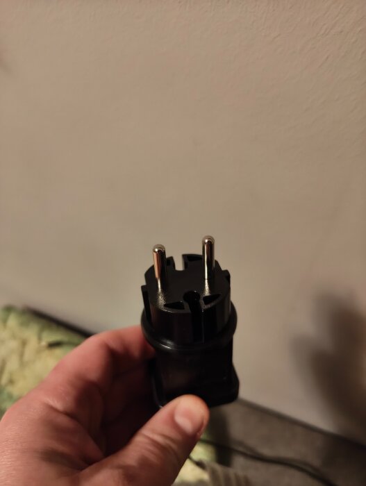 En dubbelisolerad kontakt med två stift hålls i en hand mot en beige väggbakgrund.