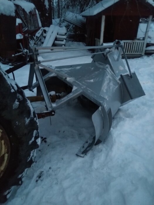 Snöplog fäst vid en traktor i en snötäckt trädgård, omgiven av några snöiga byggnader och staplade trästockar.