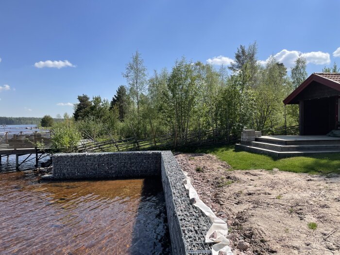 Gabioner vid en sjö, omgivna av träd och en röd byggnad, med vatten som når gabionernas botten.