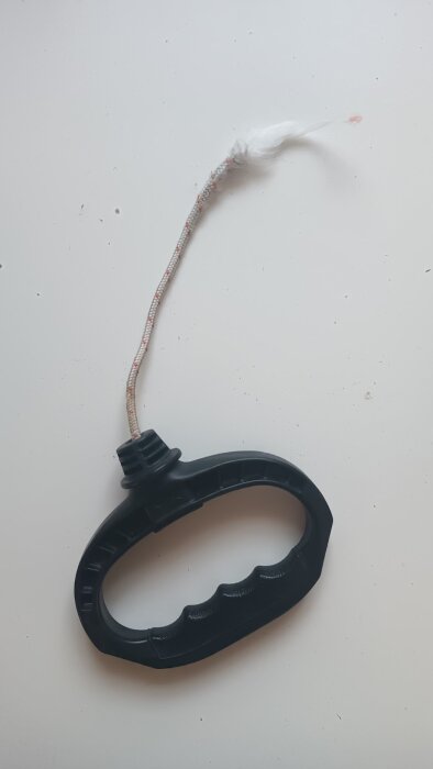 Svart plastgrepp med ett trasigt snöre fäst vid ena änden, liggande på ett ljusgrått underlag.