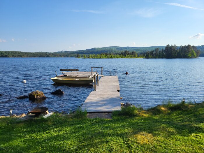 Flytbrygga med båt vid insjö, omgiven av skog och berg. En person simmar i vattnet nära bryggan under en klarblå himmel.