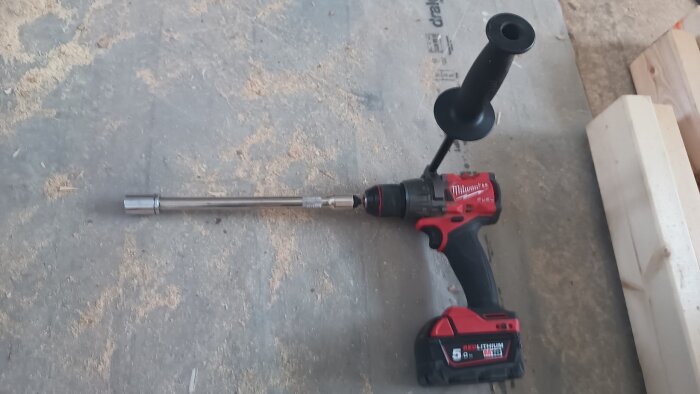En batteridriven borrmaskin med lång förlängning ligger på ett betonggolv med sågspån runt omkring.