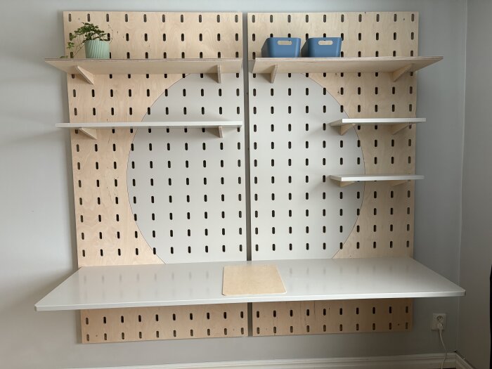 Verkstadsinspirerad pegboard av björkplywood med hyllor och skrivbord i målad MDF, en växt och två blå förvaringslådor.