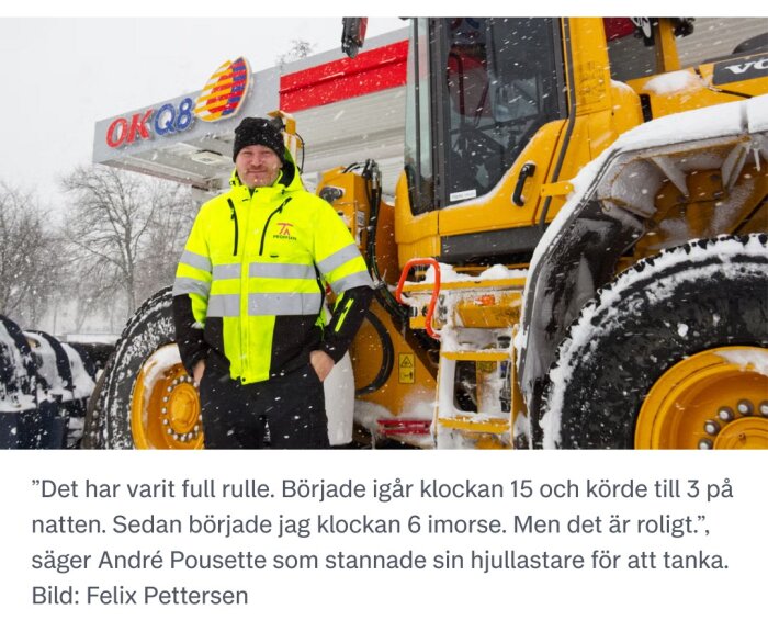 Man i gul reflexjacka står framför en stor gul hjullastare vid en OKQ8-mack, omgiven av snöfall.