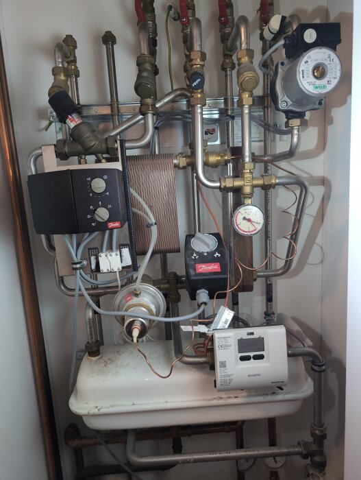 Värmepump med olika rörledningar och ventiler, inklusive en tryckmätare och Danfoss-styrenhet, installerad i ett hem.