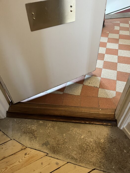 Dörröppning med tröskel, delvis synlig betong under dörren och trägolv i förgrunden. Checkerplattor på golvet i trapphuset utanför.