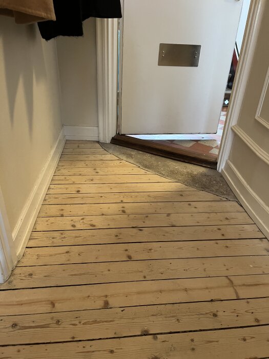 Trägolv i hall som möter betonggolv vid ytterdörren, med dörren delvis öppen mot trapphus med klinkergolv.
