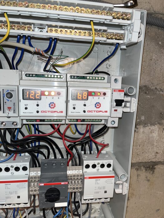 Elektrisk panel med kretsbrytare och mätare från märket Octopus, visar digitala skärmar och olika kablar uppkopplade ovanför en stenvägg.