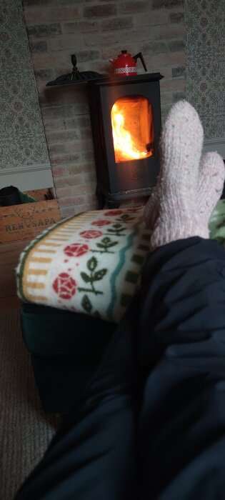 Person med rosa sockor vilar framför en braskamin med eld; tegelvägg och filt med blommönster syns i bakgrunden.