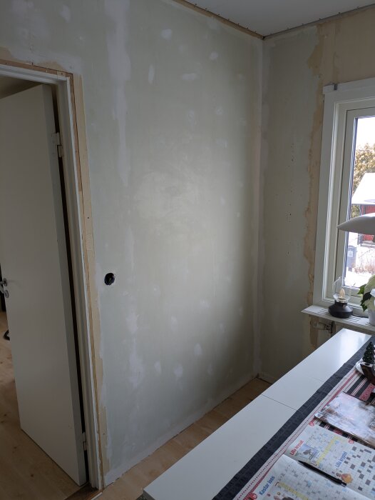 Ommålad vägg i ett rum med fönster och bord med tidningar. Under renovering med spacklade fläckar nära dörrkarm.