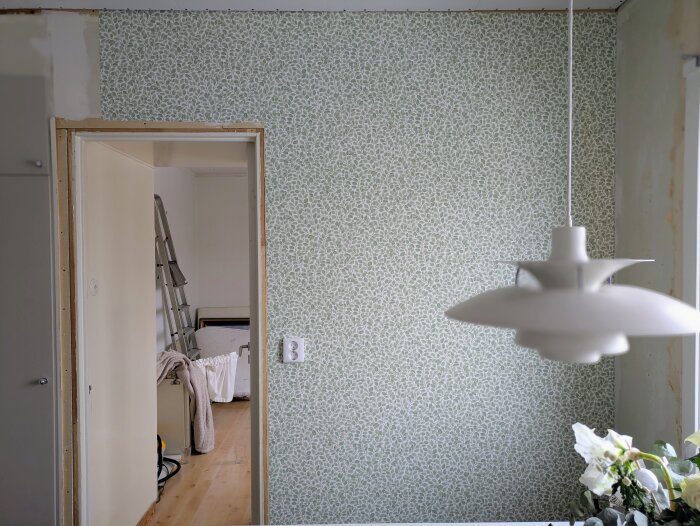 Rum under renovering med mönstrad tapet, taklampa och blommor i förgrunden. En stege och en vilandes filt syns genom dörröppningen.