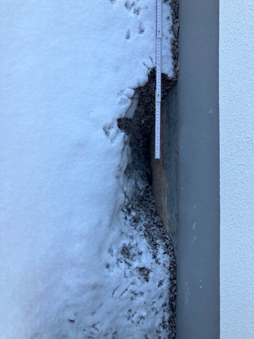 Slukhål vid husgrundsmur med snö och grus runtom, måttband visar ungefär 50 cm brett och 30 cm djupt.