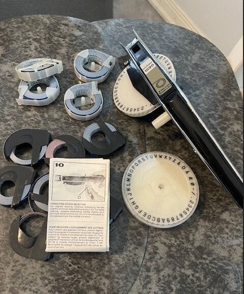 Dymo 1590 etikettskrivare med svart handtag, runda bokstavsrattar och flera märktejprullar på ett bord.