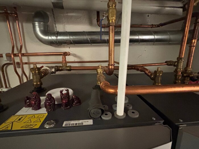 Kopparrör och ventiler ovanpå en värmepump i ett teknikrum, med små dekorationer av tre figurer på pumpen.