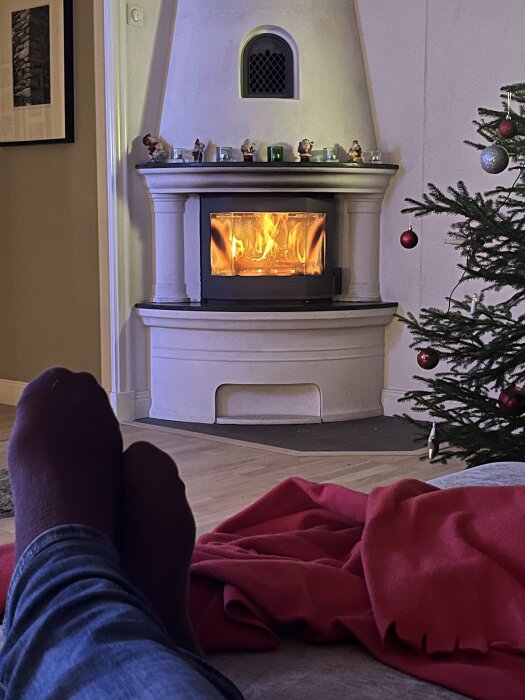 Person vilar med fötterna upp, tittar på en brasa i en kamin. Bredvid finns en dekorerad julgran och en röd filt ligger på soffan.