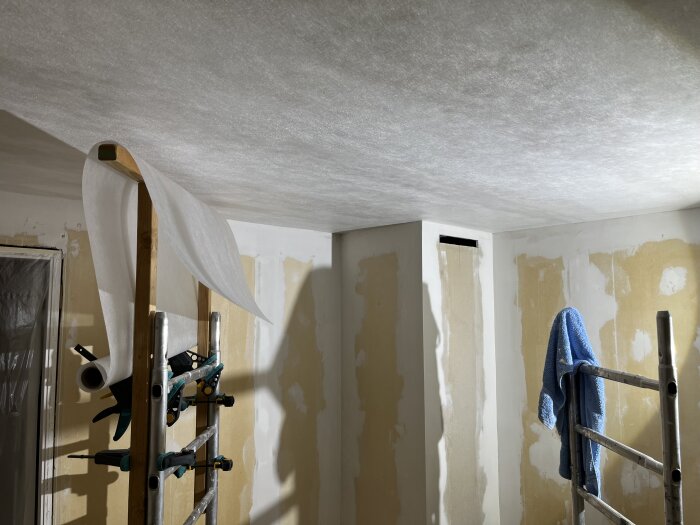 En stege med fästa verktyg och rulle av microlit i ett rum med renoverade väggar och blå handduk på en byggställning.