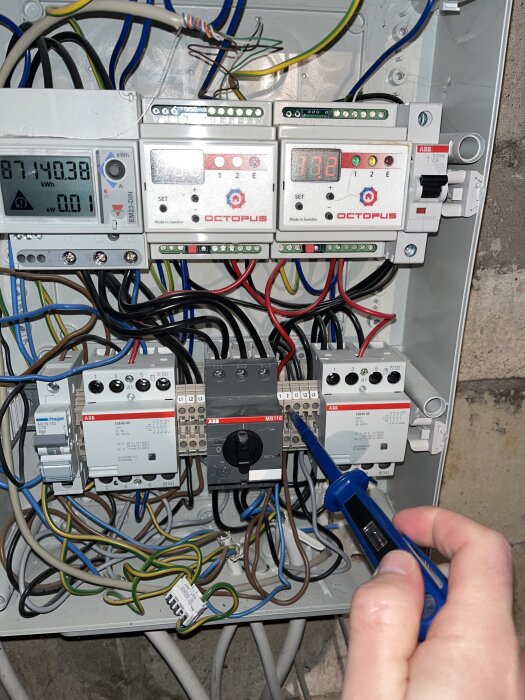 Person testar elektrisk central med testpenna, flera strömbrytare och kablar synliga.