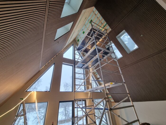Byggställning i högt rum med träpanel-tak och stora trekantiga fönster, används för pågående renovering i hem.