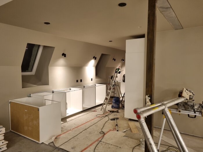 Kök under renovering med montering av vita köksskåp, stege, verktyg och byggmaterial. Utrymmet är upplyst och oavslutat.