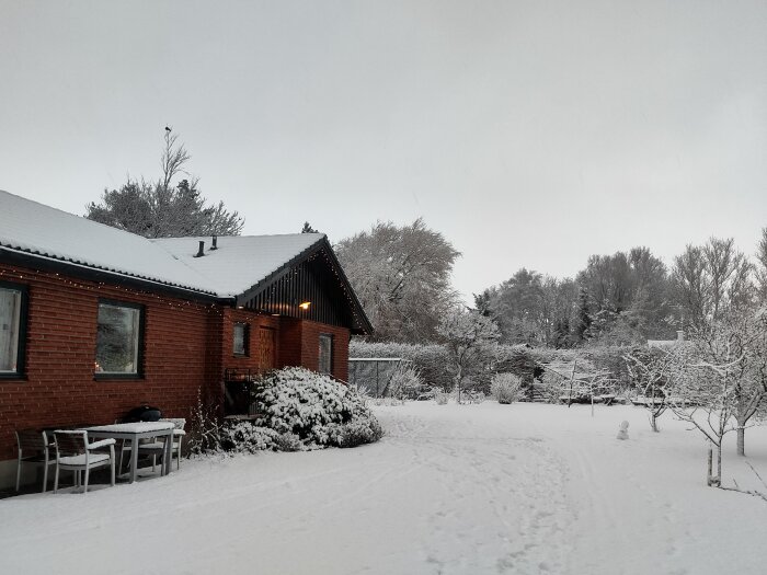 Röd tegelbyggnad omgiven av snö med buskar, träd och utemöbler täckta av snö; vinterlandskap.