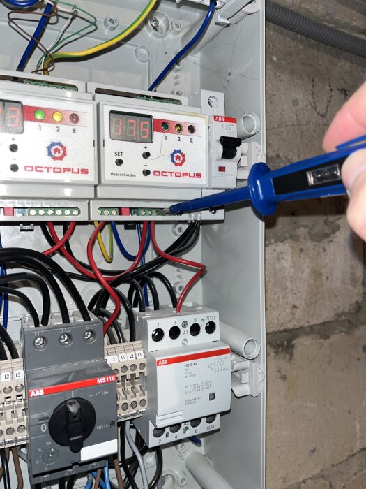 Elektrisk kopplingslåda med flera brytare och en spänningstestare riktad mot en röd kabel på höger sida.