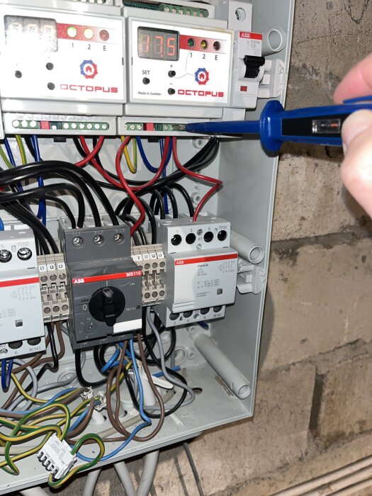 Elektrisk kopplingsbox med ledningar och mätutrustning. En person testar en röd kabel till höger med en testmejsel.