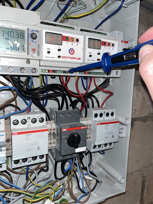 Elektriskt kopplingsskåp med olika mätinstrument, kablar och brytare. Hand håller en testmejsel vid anslutningarna.