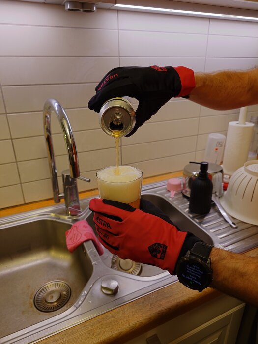 Person med röda arbetshandskar häller öl från en burk i ett glas vid en diskbänk.