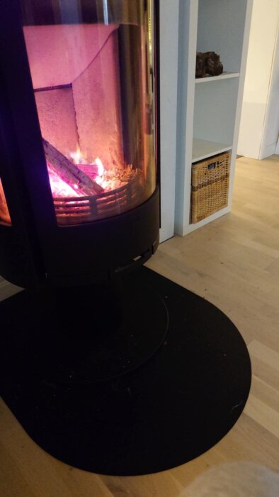 Kamin med eld brinnande inuti, omgiven av svart metall och ljus träinredning i bakgrunden.