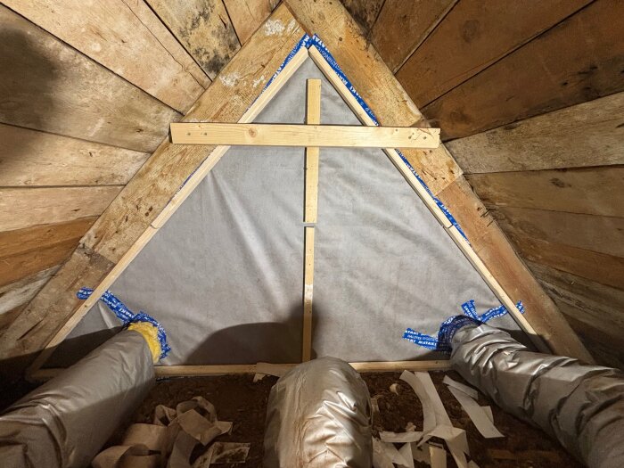 Isolering och duk installerade på en trävägg i ett loftutrymme, med blå tejp och ventilationsrör synliga.