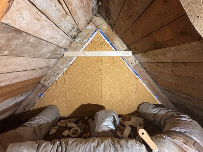 Ett vindsutrymme med nyinstallerad isolering på väggarna, omgiven av träpanel och ventilationsrör, samt verktyg och tejp synliga på golvet.