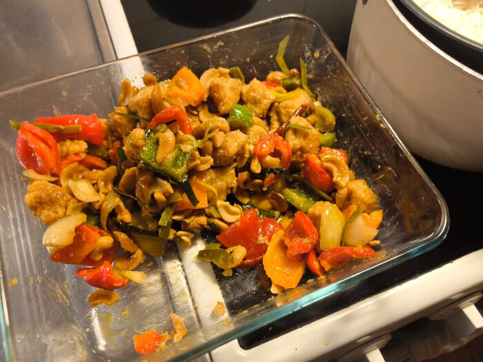 Thailändsk kycklingrätt med paprika och cashewnötter i en glasform på spis, intill en kastrull med ris.