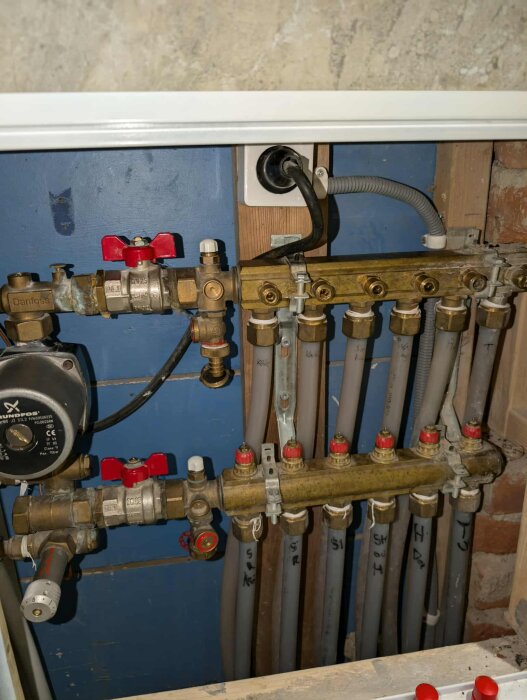 VVS-installation i ett hem med rörledningar och ventiler, bland annat en pump och kopplingar, monterade på en blå bakgrund och omgivna av trä och tegel.
