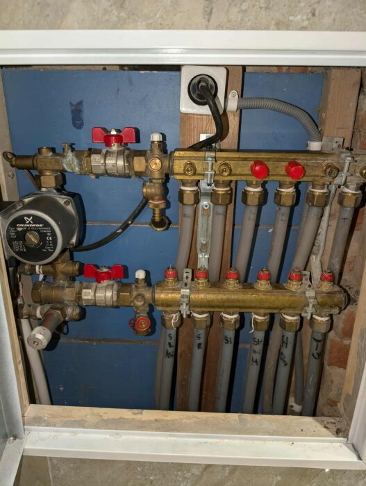Rörinstallation med flera ventiler och kopplingar, troligen för värme- eller vattensystem, monterad i en väggnisch med en elektrisk kabel och stickkontakt.