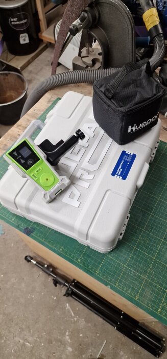 Grön och svart lasermätare på en vit Huepar-låda, med en svart väska bredvid, placerade på ett arbetsbord.