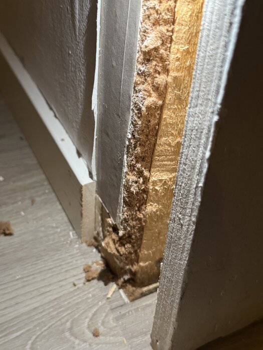 Närbild av en öppnad vägg där innanmätet och isoleringen syns, mot en trägolvyta med några träspån och damm.