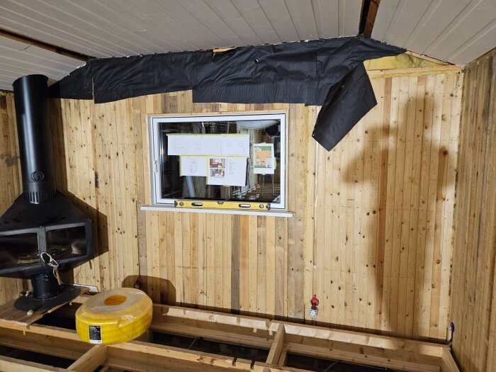 Inre vägg med träpanel, exponerad ångspärr och konstruktionsdetaljer, inklusive fönster och vedspis i vänster hörn.
