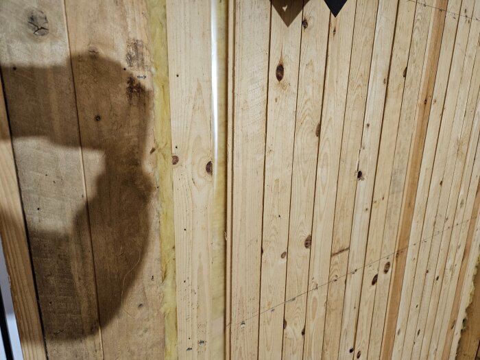 Trävägg med träpanel där ångspärr är delvis synlig bakom träpanelen. Skuggor och perforeringar i ångspärren kan ses på bilden.