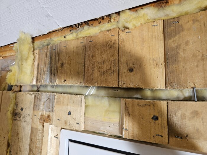 Skadad och perforerad ångspärr synlig bakom invändig träpanel, med isolering och spik, under pågående renovering i enplanshusets vägg.