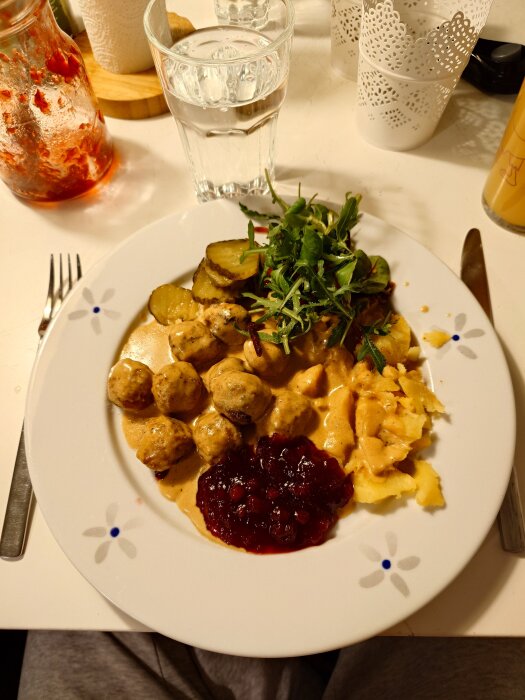 Tallrik med köttbullar, sås, mosad potatis, lingonsylt, syltgurka och sallad, serverad på ett bord med glas och servetter.