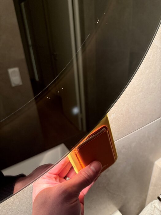 Hand håller upp något bakom en genomskinlig spegel med belysning, spegeln visar sprickor och den reflekterar ett handfat i bakgrunden.
