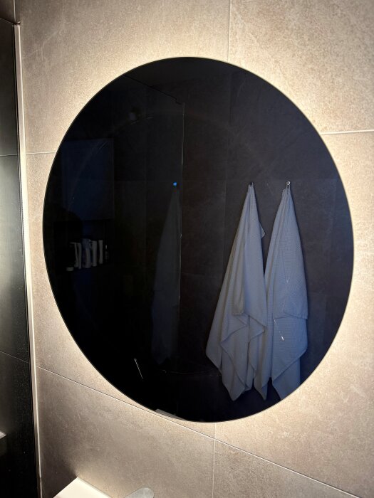 Rund spegel i badrum med synlig bakgrundsbelysning och två hängande handdukar reflekterade på kaklade väggen.