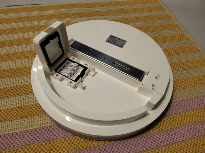 Vit rund elektrisk takarmatur med avtagbar baksida och exponerad kopplingsdosa, placerad på en randig matta i gult och lila.